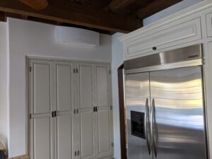 White wall mounted mini split air conditioner mounted under a dark wooden ceiling above a white wardrobe in the corner of the room.