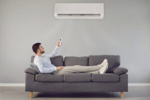 A man is lying on a couch under a wall with an air conditioner hanging on it.
